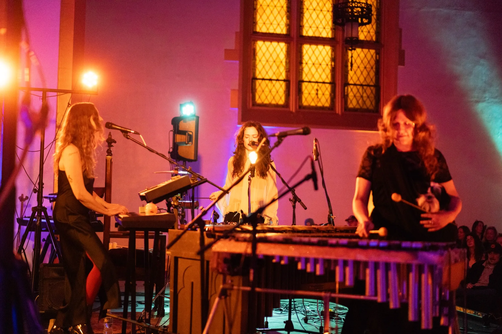 Three bandmates performing on stage under purple light: one on keyboard, one singing at the mic, one playing vibraphone with mallets.