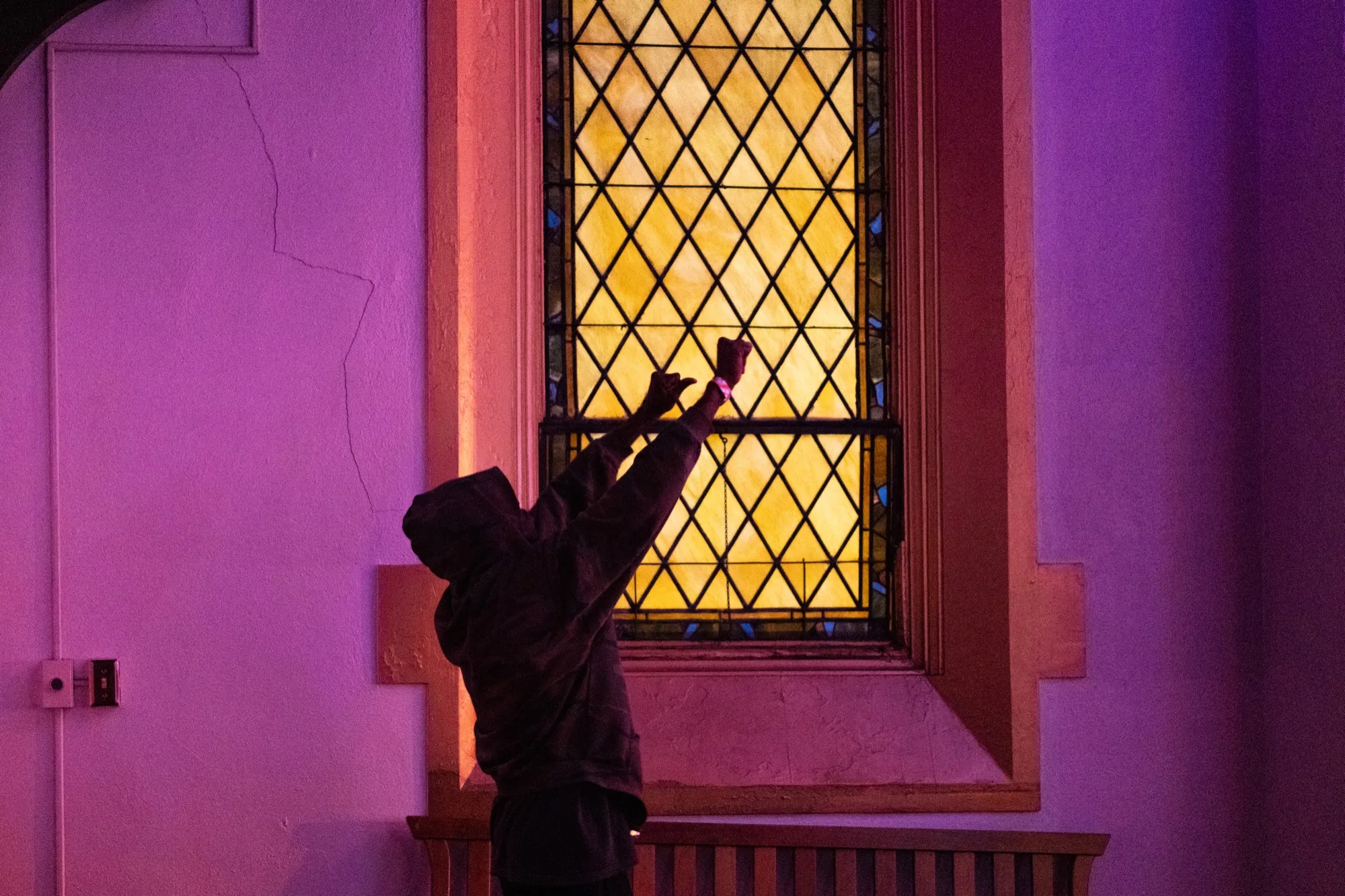 A hooded figure reaching up to touch a glowing stained glass window.