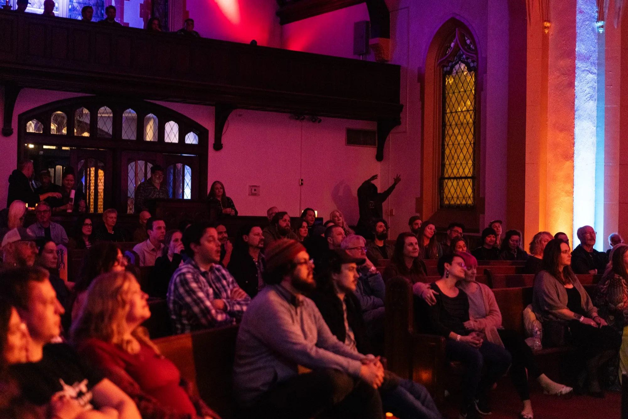 An audience seated in pews watching the show, lit in red and purple stage light.