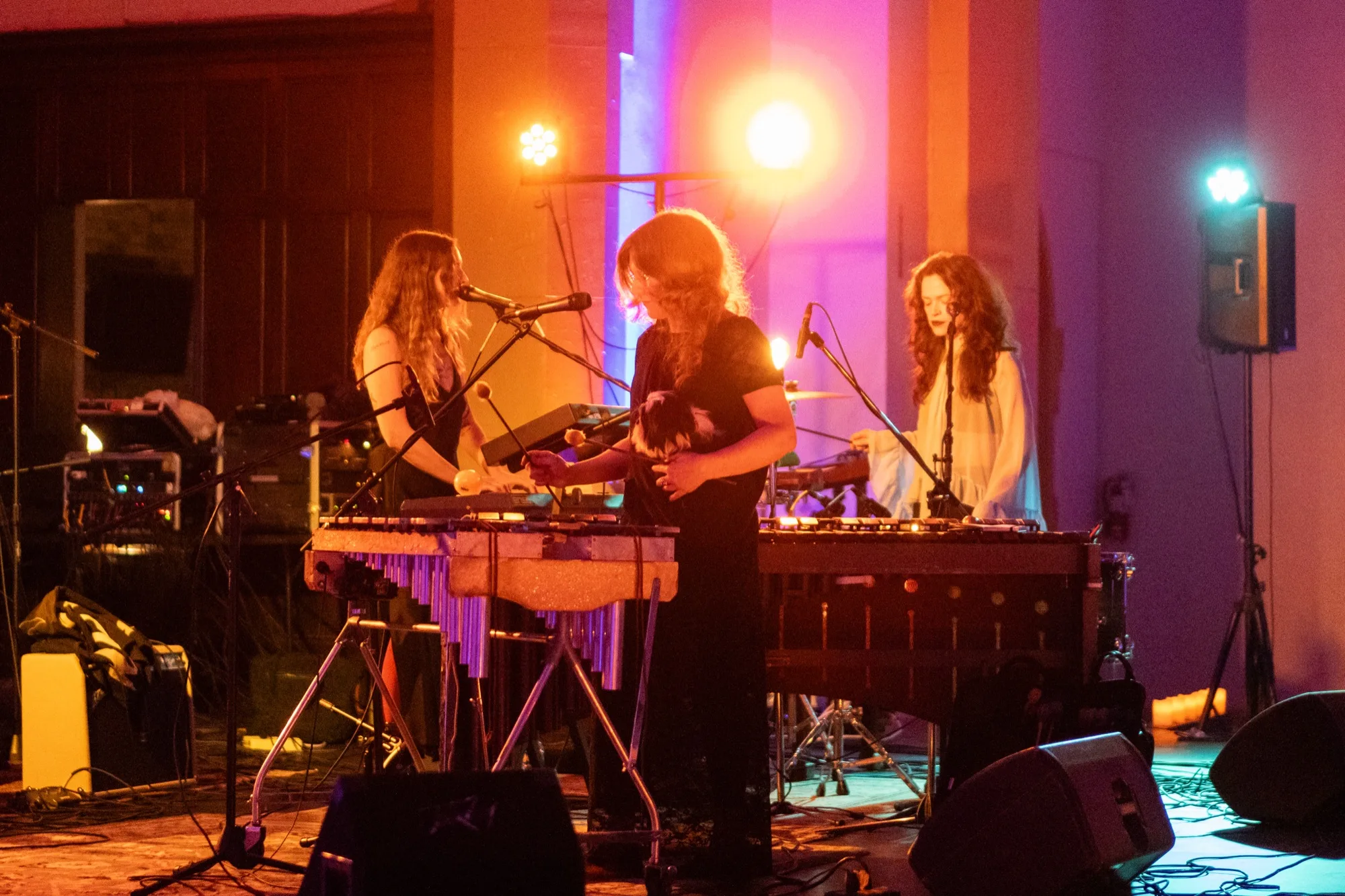 Three bandmates performing together at a row of keyboards and a vibraphone under blue and pink light.