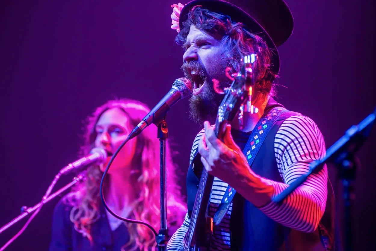 A bandmate in a hat singing and playing guitar, another bandmate singing behind him, both lit in purple stage light.