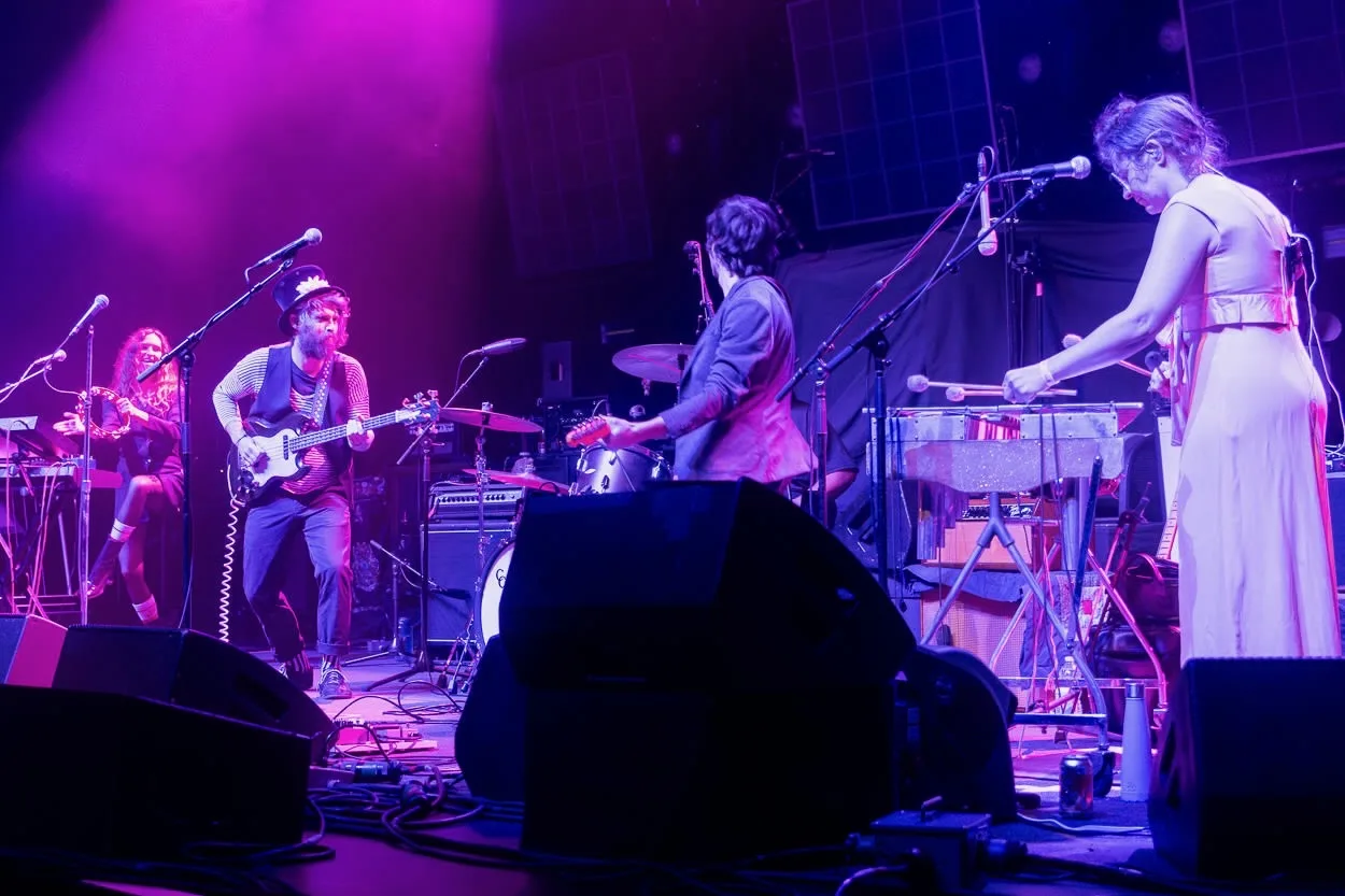 Wide shot of the full band performing on stage under purple light, a bandmate in a long dress playing keyboard.