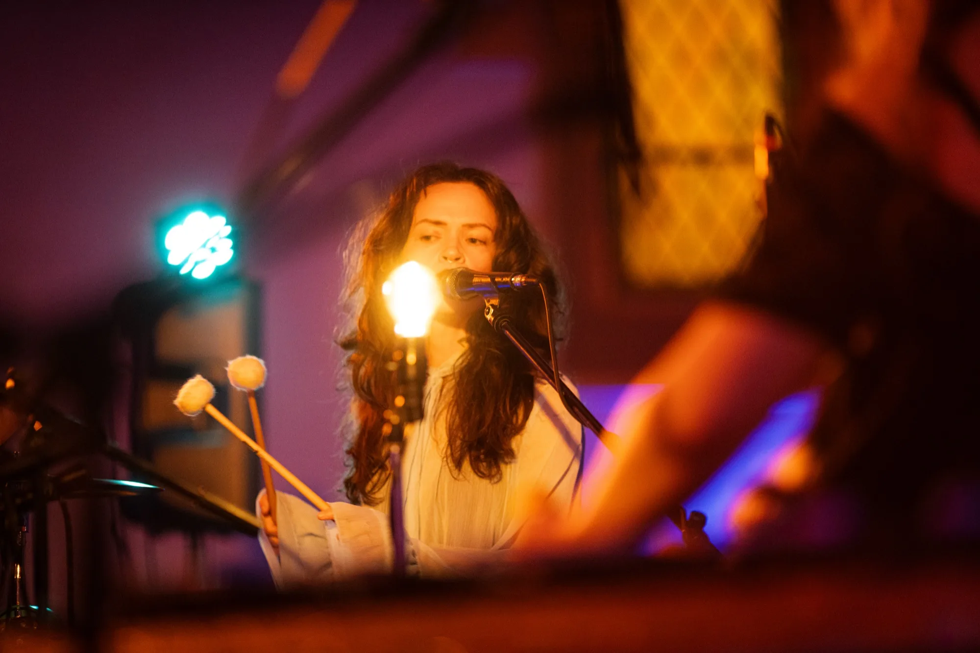 Close-up of Regan Heckscher playing mallets on a vibraphone and singing into a mic, lit by a small lamp in a purple room.